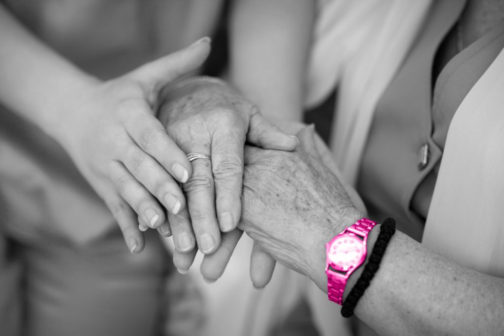 Image shows an elderly persons hands held by a younger persons. The elderly person wears a watch and bracelet around their wrist.