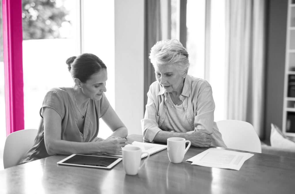 Image shows a middle-aged woman sat at table with her mother. They are both looking at a piece of paper on the table with mugs of tea. They sit in front of a floor to ceiling window.