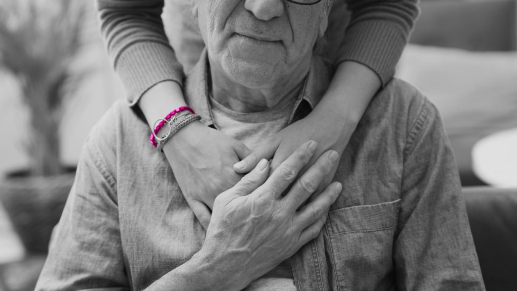 Image shows older man sat with younger woman's arms wrapped around his shoulders.