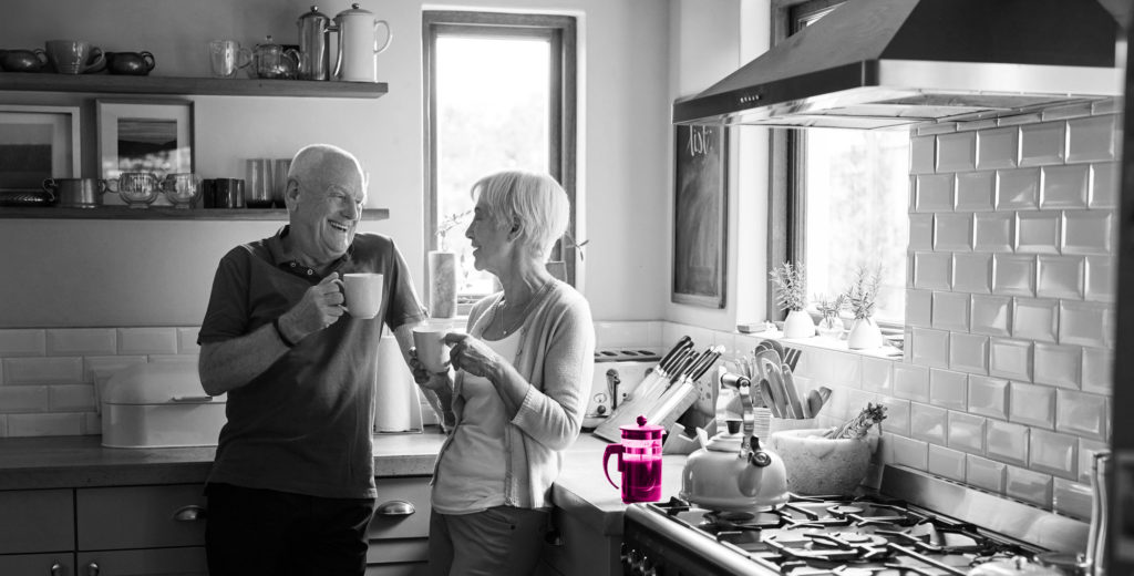 Image shows elderly couple drinking coffee in their kitchen. They are stood next to a stove with a kettle boiling on it and a cafetiere brewing coffee on the kitchen side.